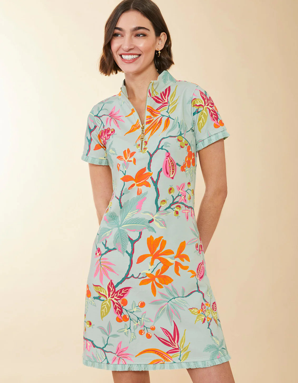 A woman wearing a short-sleeve collared dress with a colorful tropical floral print on a light mint green base, posed against a solid beige studio background.