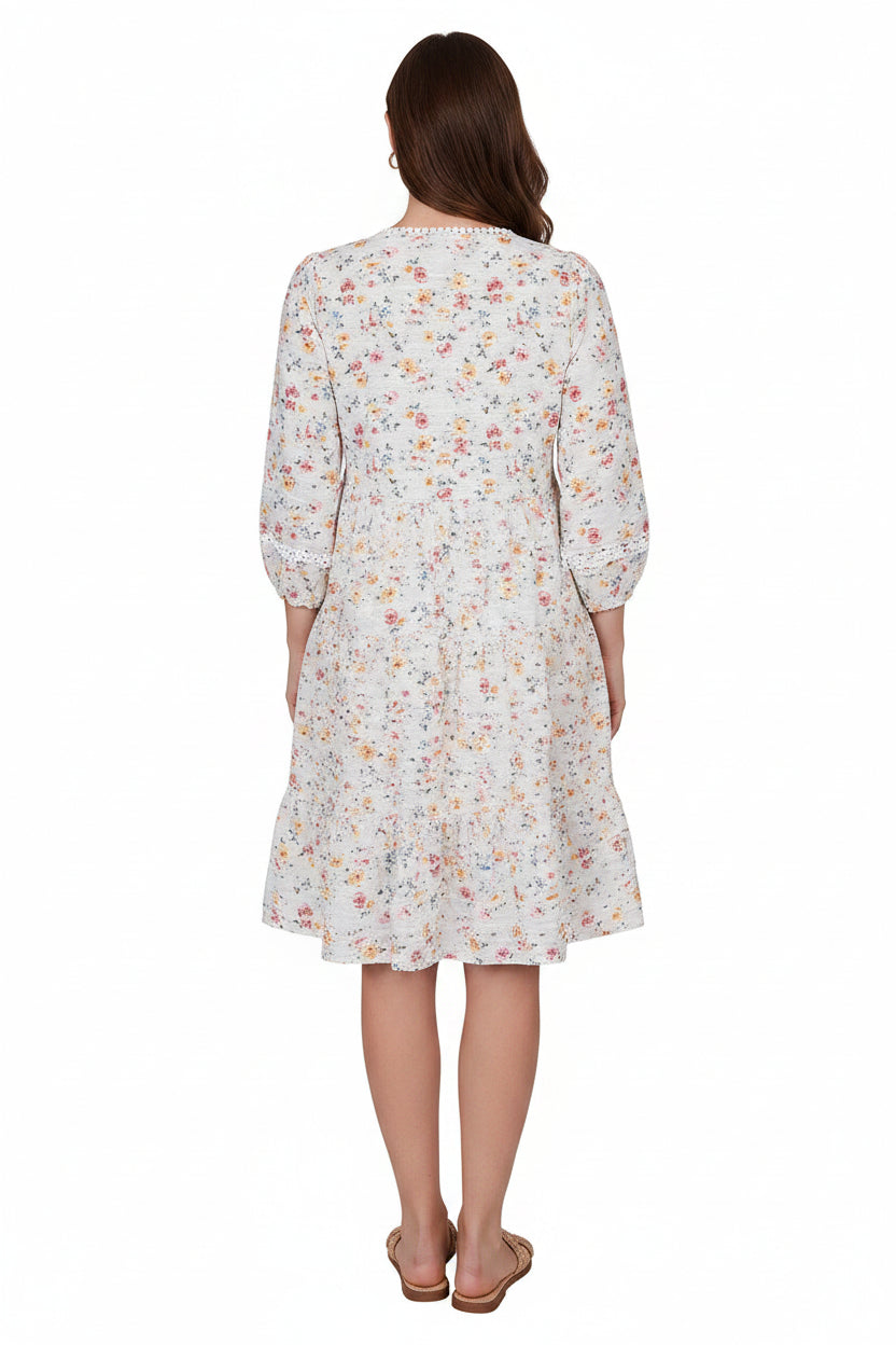 A woman modeling a knee-length white dress with a colorful floral print, featuring three-quarter sleeves and a V-neckline, set against a plain white background.