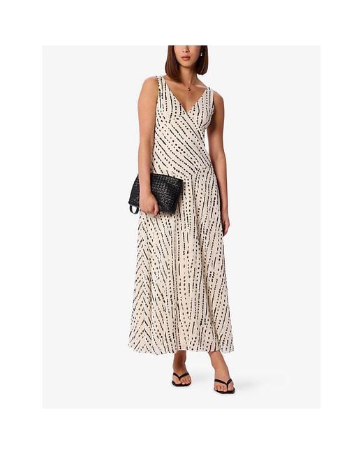 A woman models a sleeveless, V-neck maxi dress with a black abstract vertical pattern on a cream background. She is holding a small black woven clutch and wearing black sandals, set against a plain light gray studio background.