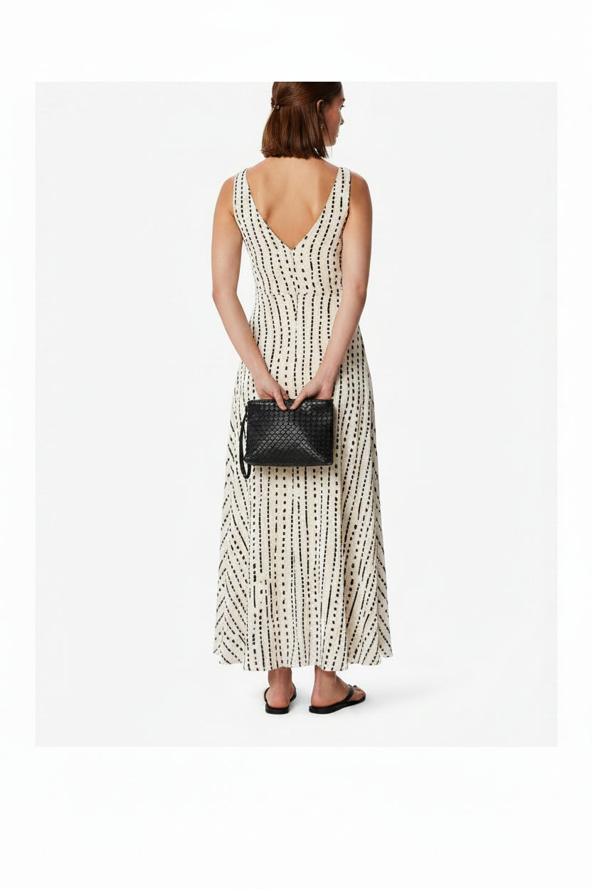 A woman models a sleeveless, V-neck maxi dress with a black abstract vertical pattern on a cream background. She is holding a small black woven clutch and wearing black sandals, set against a plain light gray studio background.
