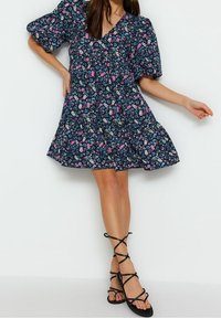 A woman wearing a navy blue floral puff-sleeve mini dress with a V-neckline, paired with black lace-up flat sandals, against a plain white background.