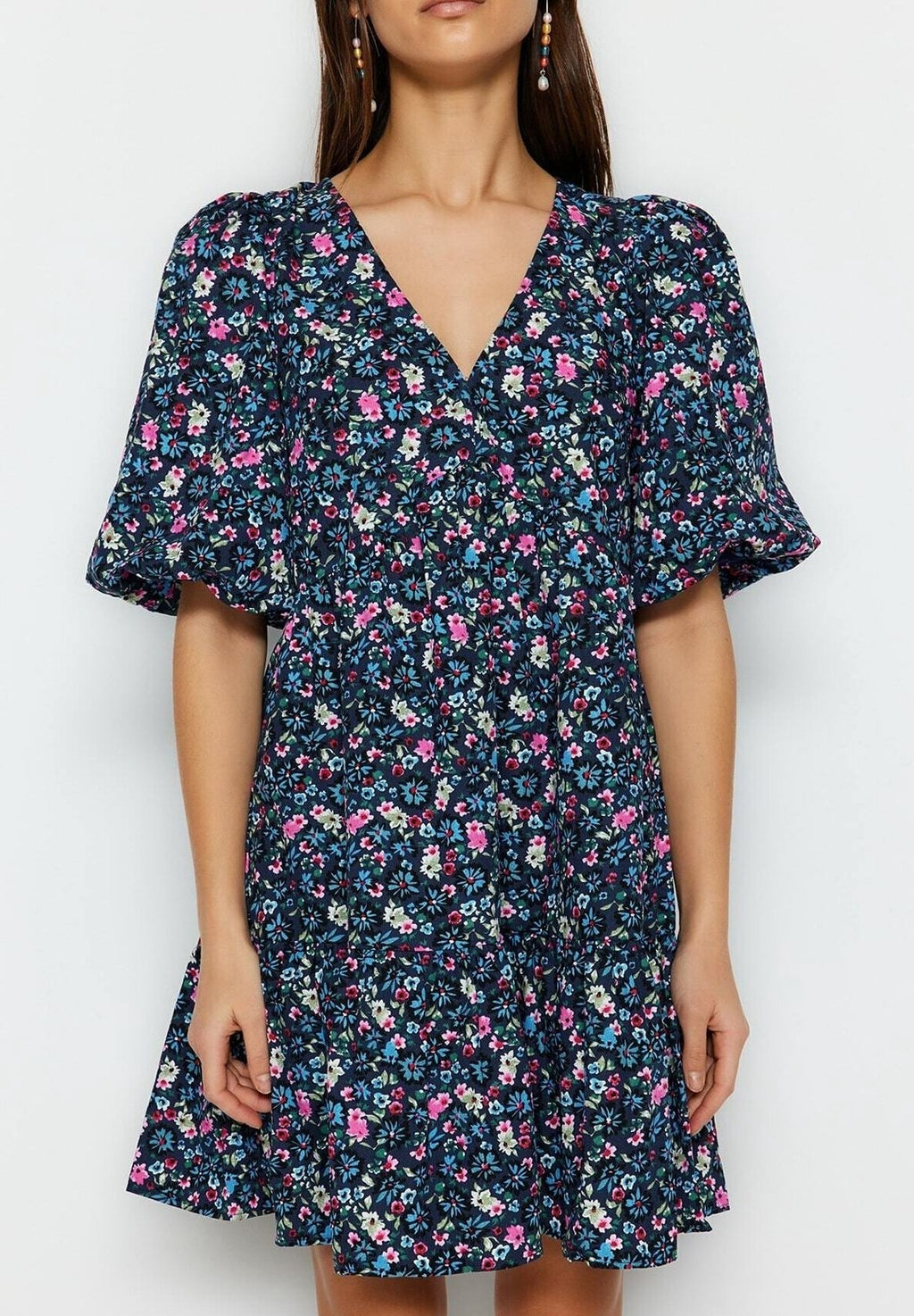 A woman wearing a short-sleeve V-neck dress with a dark navy blue background and a dense pink, purple, and white floral print. The dress features puff sleeves and a flared skirt, shown against a plain light gray background.