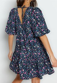 A woman wearing a short-sleeve floral dress with a tie-back neckline, shown from the back against a plain light background.