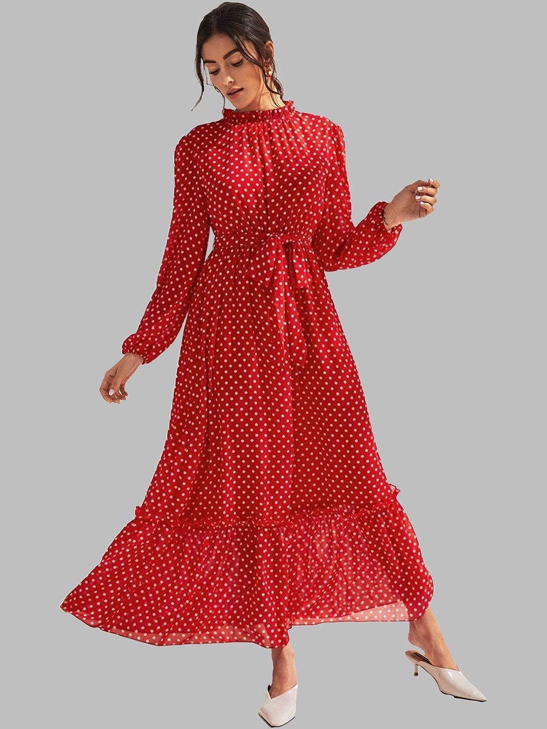 A woman modeling a long-sleeve red maxi dress with a white polka dot pattern, featuring a high ruffled neckline, a matching fabric tie belt at the waist, and a tiered skirt, set against a solid gray background.
