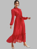 A woman modeling a long-sleeve red maxi dress with a white polka dot pattern, featuring a high ruffled collar, a defined waist, and a tiered ruffle hem, paired with beige pointed-toe mule heels against a solid gray background.